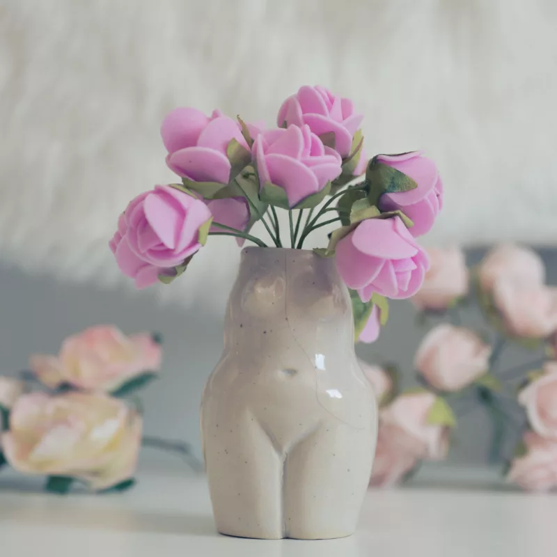 Présentation d'une fiole en céramique aux formes d'une femme, contenant des fleurs roses.