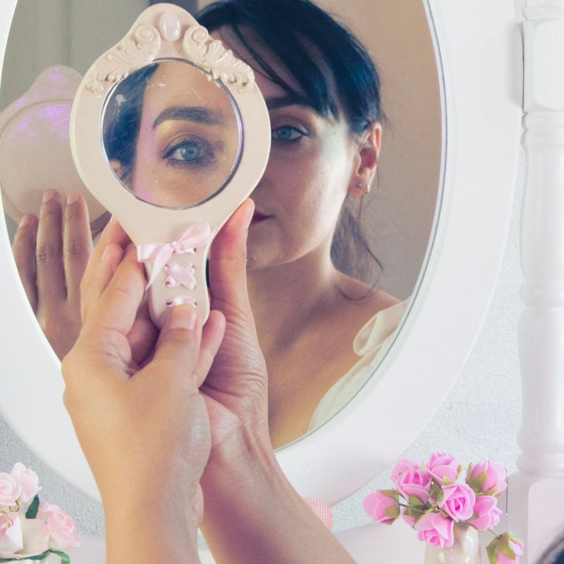 femme tenant un Mini miroir en céramique, orné d'un lacet de satin rose, avec quelques fleurs rose autour. on aperçoit sont reflet dedans
