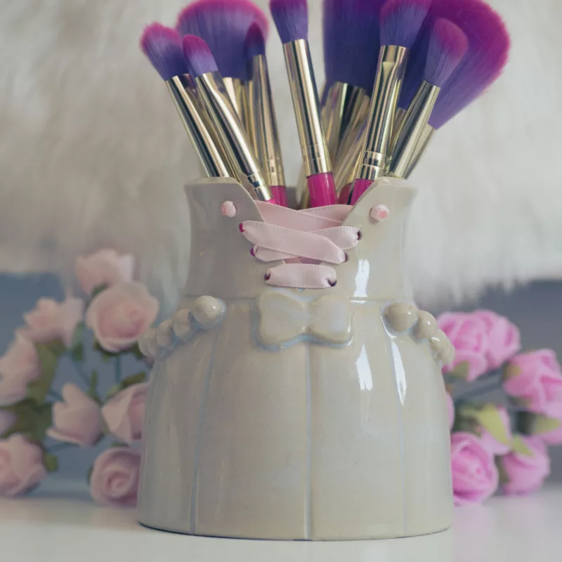 Petit vase en grès, orné d'un lacet de satin rose, contenant des pinceaux à maquillage roses et violets, avec des fleurs roses de chaque côté du pot.
